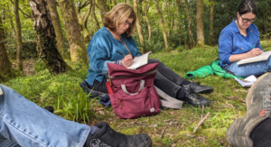 Individuals writing in notebooks sitting on grass in a forest setting.