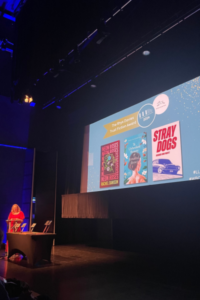 A person on stage with a screen behind them showing books.