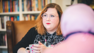 A person in a black and white dress sitting at a table talking to another person wearing a pink headscarf