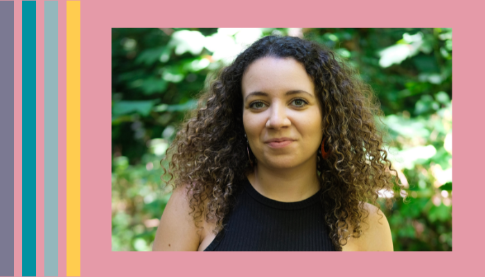 a woman with dark curly hair wearing a black top stands in front of some trees